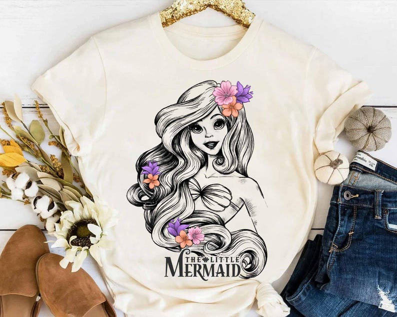 May include: Cream-colored t-shirt featuring a black and white illustration of a mermaid with long flowing hair adorned with purple and orange flowers. The text "THE LITTLE MERMAID" is printed below the illustration.