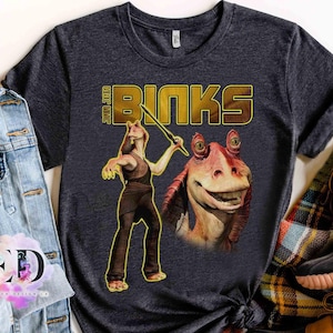 May include: Dark gray t-shirt featuring a graphic of Jar Jar Binks from Star Wars. The design includes the text "JAR JAR BINKS" in gold lettering, and an illustration of the character holding a weapon. The shirt is a crew neck.