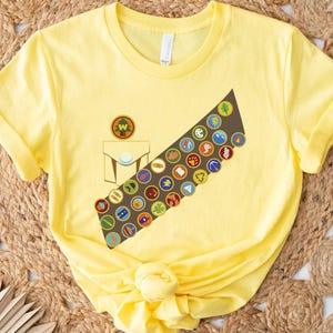 May include: A yellow t-shirt featuring a brown sash adorned with colorful merit badges. The shirt also displays a pocket design with a circular emblem. The design is reminiscent of scouting or youth organization attire.