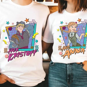 Pode incluir: T-shirts brancas com ilustra&ccedil;&otilde;es de desenhos animados de Kristoff e Anna de Frozen. A camisa de Kristoff tem o texto "Her Kristoff" e a camisa de Anna tem o texto "His Anna". Ambas as camisas t&ecirc;m um estilo retro dos anos 80.