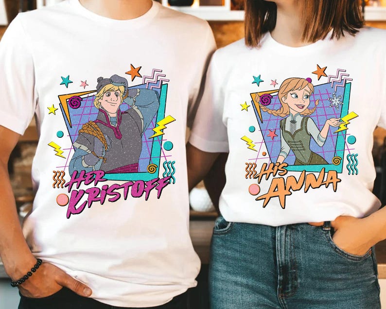 Pode incluir: T-shirts brancas com ilustra&ccedil;&otilde;es de desenhos animados de Kristoff e Anna de Frozen. A camisa de Kristoff tem o texto "Her Kristoff" e a camisa de Anna tem o texto "His Anna". Ambas as camisas t&ecirc;m um estilo retro dos anos 80.