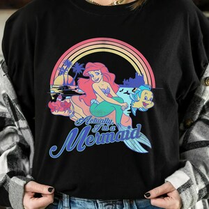 Pode incluir: Camiseta preta com um gr&aacute;fico colorido de uma sereia de cabelo ruivo, sentada numa rocha com um peixe. O design inclui um arco-&iacute;ris, palmeiras e o texto "Actually I'm a Mermaid."