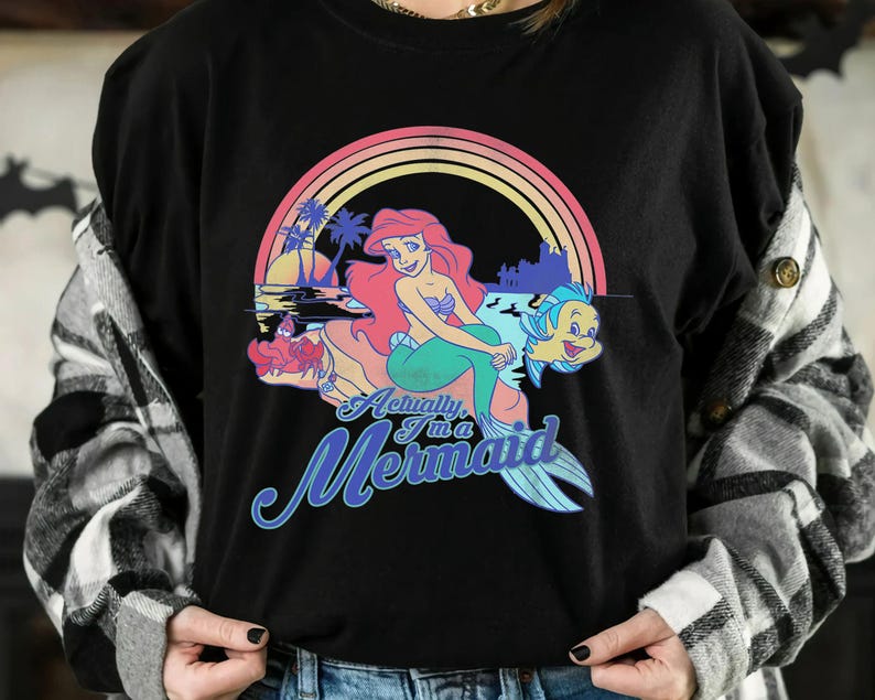 Pode incluir: Camiseta preta com um gr&aacute;fico colorido de uma sereia de cabelo ruivo, sentada numa rocha com um peixe. O design inclui um arco-&iacute;ris, palmeiras e o texto "Actually I'm a Mermaid."