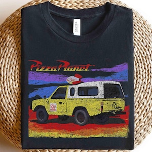 Może przedstawiać: Czarna koszulka z grafiką ciężar&oacute;wki Pizza Planet z Toy Story. Ż&oacute;łta ciężar&oacute;wka ma białą kabinę i czerwono-białe detale. Logo Pizza Planet znajduje się nad ciężar&oacute;wką, z kolorowym tłem zachodu słońca.