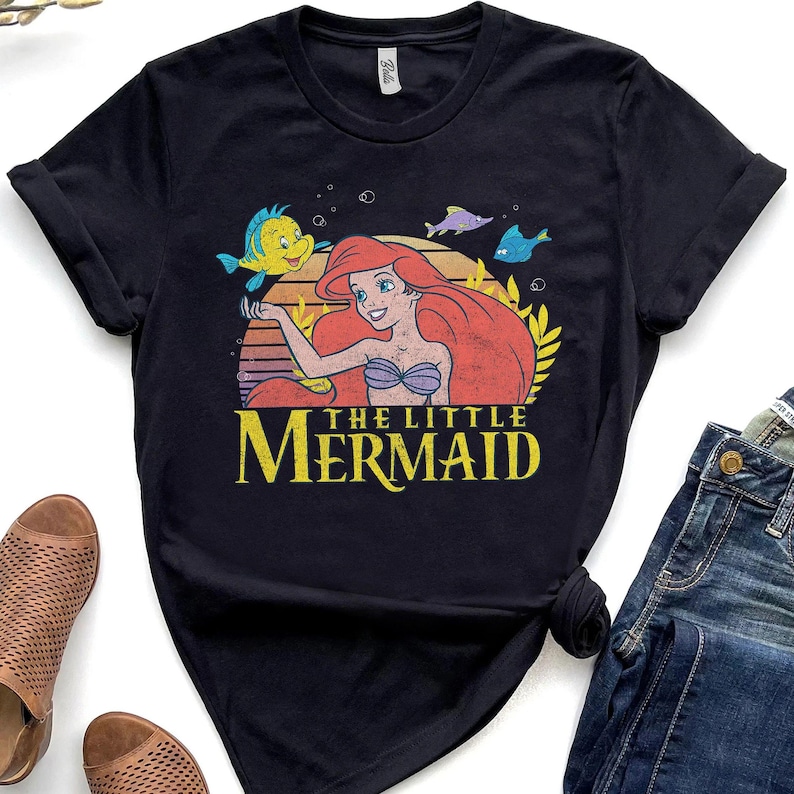May include: Black t-shirt featuring a graphic of Ariel from The Little Mermaid. The design includes Ariel with red hair, a purple seashell bra, and the text "THE LITTLE MERMAID" in yellow. The shirt also features Flounder and other sea creatures.