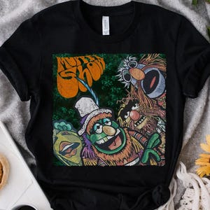 May include: Black t-shirt featuring a colorful graphic of Muppet Show characters. The design includes Kermit the Frog, Fozzie Bear, and other Muppets, with the show's logo in orange. The shirt is made of a soft material.