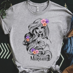 May include: A gray t-shirt featuring a black and white illustration of a mermaid with long flowing hair adorned with pink and orange flowers. The text "THE LITTLE MERMAID" is printed below the illustration.