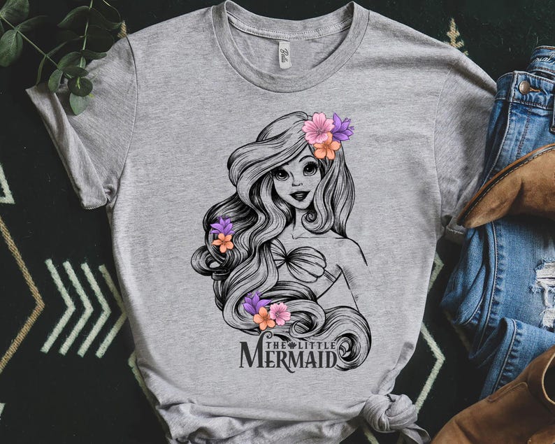 May include: A gray t-shirt featuring a black and white illustration of a mermaid with long flowing hair adorned with pink and orange flowers. The text "THE LITTLE MERMAID" is printed below the illustration.