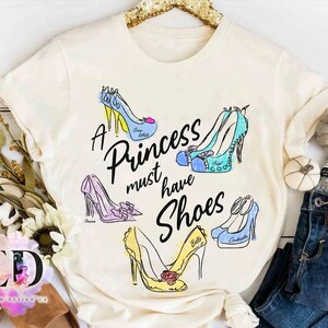 May include: A cream-colored t-shirt featuring the black text A Princess must have Shoes surrounded by five illustrated high-heeled shoes in blue, teal, purple, and yellow, each labeled with names including Snow White, Ariel, Aurora, Belle, and Cinderella.