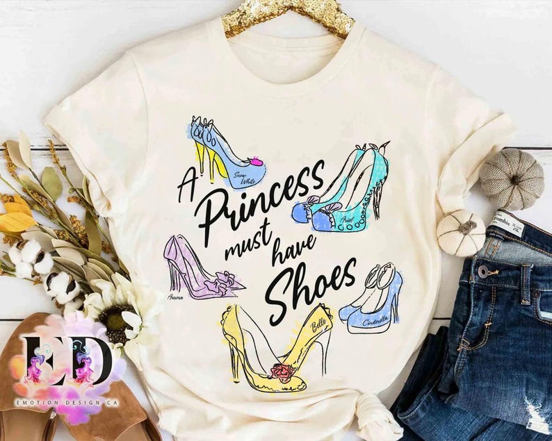 May include: A cream-colored t-shirt featuring the black text A Princess must have Shoes surrounded by five illustrated high-heeled shoes in blue, teal, purple, and yellow, each labeled with names including Snow White, Ariel, Aurora, Belle, and Cinderella.