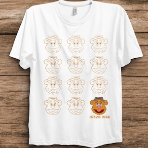 May include: A white short-sleeved t-shirt featuring a grid of eleven light orange outline illustrations of Fozzie Bear's face, with one fully colored Fozzie Bear head in the bottom right corner above the orange text FOZZIE BEAR.