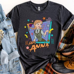 Pode incluir: T-shirt cinzento escuro com uma imagem de desenho animado de Anna de Frozen, e o texto "His Anna" em laranja e amarelo. O design inclui formas geom&eacute;tricas e raios. A camisa &eacute; estilizada com uma jaqueta jeans e botas.