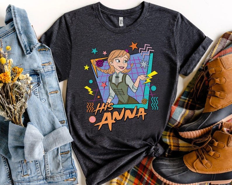 Pode incluir: T-shirt cinzento escuro com uma imagem de desenho animado de Anna de Frozen, e o texto "His Anna" em laranja e amarelo. O design inclui formas geom&eacute;tricas e raios. A camisa &eacute; estilizada com uma jaqueta jeans e botas.