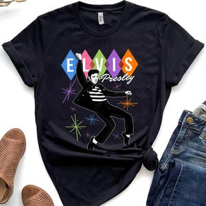 May include: Black t-shirt featuring a graphic of Elvis Presley in a dynamic pose. The design includes the name "Elvis Presley" and colorful diamond shapes. The shirt is paired with blue jeans and brown shoes.