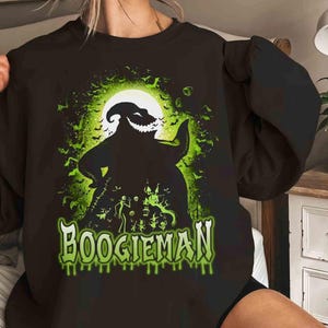 Pode incluir: Camisola preta com um gr&aacute;fico verde e preto do Boogieman de O Estranho Mundo de Jack. O design apresenta a silhueta da personagem contra uma lua cheia, com "BOOGIEMAN" em verde com letras gotejantes.