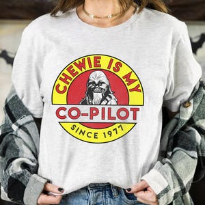 May include: A light grey t-shirt with a circular graphic. The graphic reads "CHEWIE IS MY CO-PILOT SINCE 1977" and features an image of Chewbacca in a red and yellow circle.