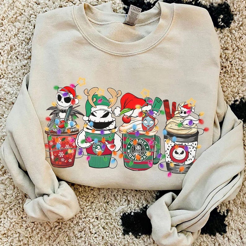 May include: Beige crewneck sweatshirt featuring a festive design of four Nightmare Before Christmas characters in coffee cup designs, each adorned with Christmas lights and holiday hats. The design includes Jack Skellington, Zero, and other characters.