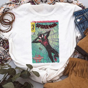 Puede incluir: Camiseta blanca con un dise&ntilde;o de portada de c&oacute;mic de Spider-Man. El gr&aacute;fico presenta a Spider-Man en una telara&ntilde;a, en rojo, negro y verde. La camiseta se combina con vaqueros azules y botas marrones con flecos.