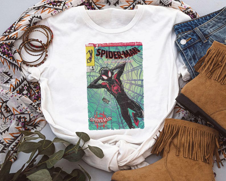 Puede incluir: Camiseta blanca con un dise&ntilde;o de portada de c&oacute;mic de Spider-Man. El gr&aacute;fico presenta a Spider-Man en una telara&ntilde;a, en rojo, negro y verde. La camiseta se combina con vaqueros azules y botas marrones con flecos.