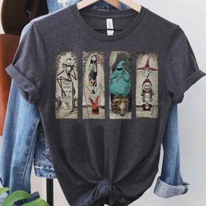 Puede incluir: Camiseta gris oscuro con cuatro paneles rectangulares con personajes de Pesadilla antes de Navidad. Los paneles incluyen a Jack Skellington, Sally, Oogie Boogie y un personaje con sombrero de bruja. La camiseta est&aacute; colgada en una percha de madera.