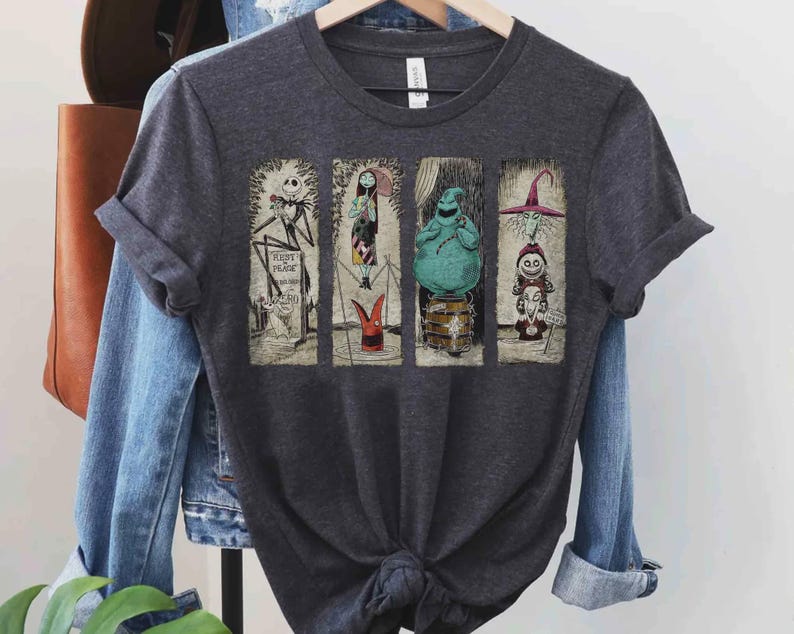 Puede incluir: Camiseta gris oscuro con cuatro paneles rectangulares con personajes de Pesadilla antes de Navidad. Los paneles incluyen a Jack Skellington, Sally, Oogie Boogie y un personaje con sombrero de bruja. La camiseta est&aacute; colgada en una percha de madera.