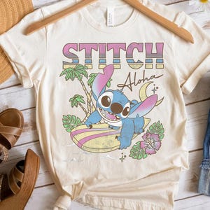 K&ouml;nnte beinhalten: Cremefarbenes T-Shirt mit dem Wort "STITCH" in Rosa, Blau und Gelb, darunter "Aloha". Eine blaue Cartoonfigur surft auf einem Surfbrett, mit Palmen und einer Blume. Das T-Shirt h&auml;ngt an einem Holzb&uuml;gel.