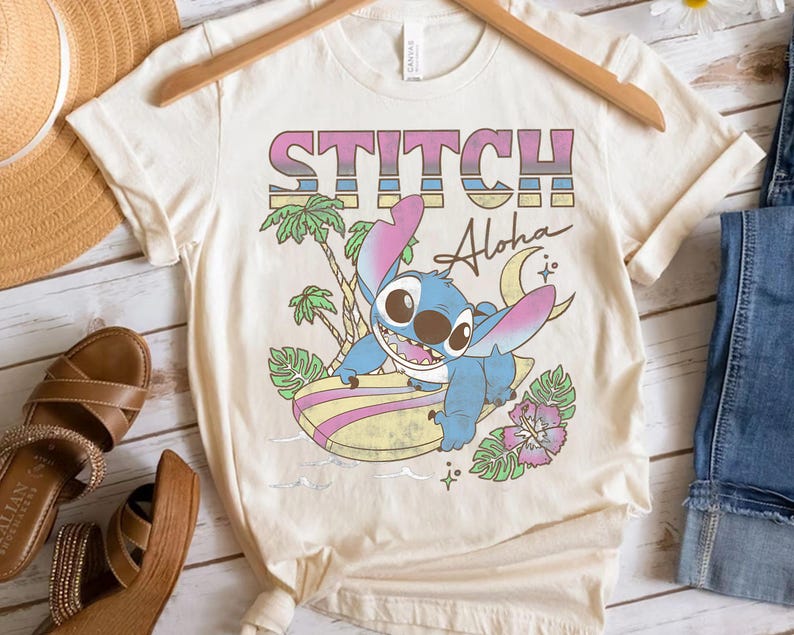 K&ouml;nnte beinhalten: Cremefarbenes T-Shirt mit dem Wort "STITCH" in Rosa, Blau und Gelb, darunter "Aloha". Eine blaue Cartoonfigur surft auf einem Surfbrett, mit Palmen und einer Blume. Das T-Shirt h&auml;ngt an einem Holzb&uuml;gel.