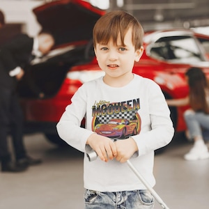 May include: A young child wearing a white long-sleeved shirt with a graphic of Lightning McQueen from the movie Cars. The shirt has the text "Lightning McQueen" above the car image. The child is holding a metal object.