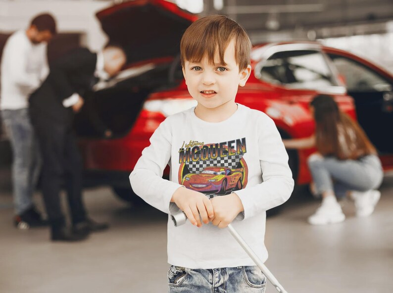 May include: A young child wearing a white long-sleeved shirt with a graphic of Lightning McQueen from the movie Cars. The shirt has the text "Lightning McQueen" above the car image. The child is holding a metal object.