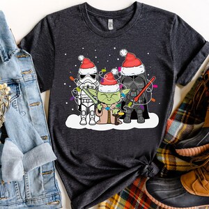May include: Dark gray t-shirt featuring a festive design with Star Wars characters. Stormtrooper, Yoda, and Darth Vader are depicted wearing Santa hats and holding Christmas lights. The shirt is displayed with a denim jacket and brown boots.