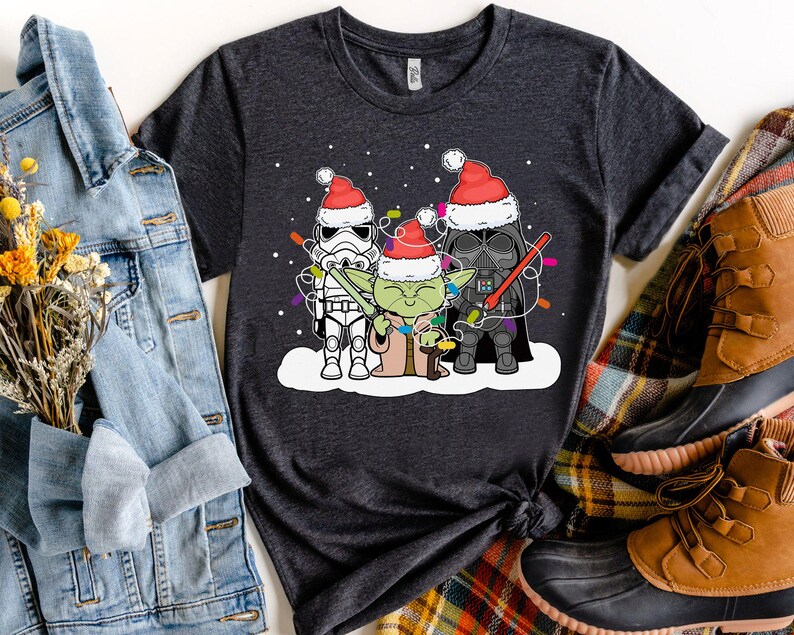 May include: Dark gray t-shirt featuring a festive design with Star Wars characters. Stormtrooper, Yoda, and Darth Vader are depicted wearing Santa hats and holding Christmas lights. The shirt is displayed with a denim jacket and brown boots.