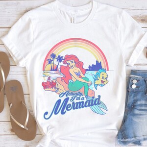 Pode incluir: Camiseta branca com um desenho gr&aacute;fico colorido de uma sereia com cabelo ruivo e cauda verde, com o texto "Actually, I'm a Mermaid". O design inclui um arco-&iacute;ris, palmeiras e um castelo. Tamb&eacute;m s&atilde;o vis&iacute;veis chinelos castanhos e cal&ccedil;&otilde;es de ganga.