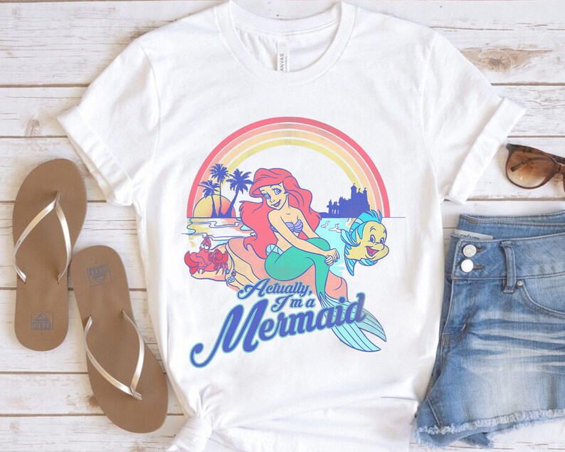 Pode incluir: Camiseta branca com um desenho gr&aacute;fico colorido de uma sereia com cabelo ruivo e cauda verde, com o texto "Actually, I'm a Mermaid". O design inclui um arco-&iacute;ris, palmeiras e um castelo. Tamb&eacute;m s&atilde;o vis&iacute;veis chinelos castanhos e cal&ccedil;&otilde;es de ganga.