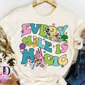 May include: Cream-colored t-shirt with the text "Every Mile is Magic" in colorful, retro-style lettering. The design includes cartoon characters like Mickey and Minnie Mouse, and a castle.
