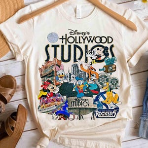 Puede incluir: Camiseta color crema con un gráfico colorido de los personajes de Disney's Hollywood Studios, incluyendo Mickey Mouse y Donald Duck. El diseño incluye el logotipo del parque y elementos relacionados con el cine. La camiseta se muestra en una percha de madera.