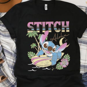 K&ouml;nnte beinhalten: Schwarzes T-Shirt mit einer Cartoon-Illustration von Stitch beim Surfen. Das Design enth&auml;lt den Text "STITCH Aloha" in einer Retro-Schrift, Palmen und einen Mond. Die Grafik ist in Blau-, Rosa- und Gelbt&ouml;nen gehalten.