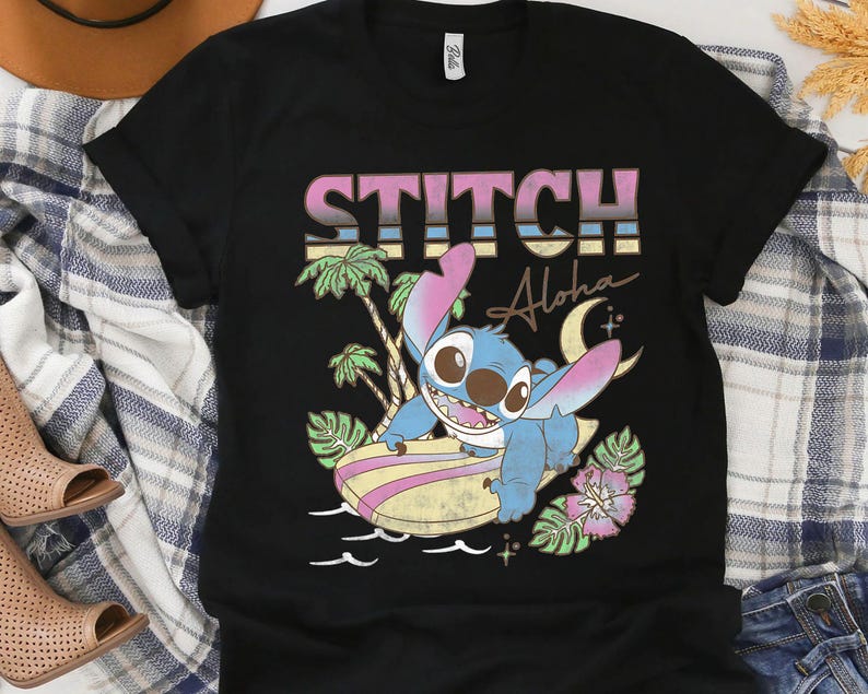 K&ouml;nnte beinhalten: Schwarzes T-Shirt mit einer Cartoon-Illustration von Stitch beim Surfen. Das Design enth&auml;lt den Text "STITCH Aloha" in einer Retro-Schrift, Palmen und einen Mond. Die Grafik ist in Blau-, Rosa- und Gelbt&ouml;nen gehalten.