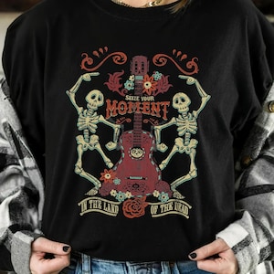 May include: Black long-sleeve shirt with a colorful graphic featuring a guitar, skeletons, and floral elements. The text reads "Seize Your Moment" and "In The Land Of The Dead". The design has a Day of the Dead theme.