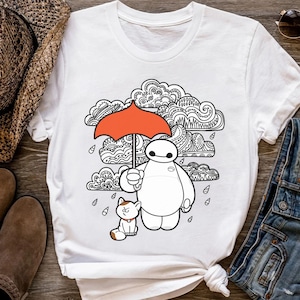 May include: White t-shirt featuring a cartoon illustration of a white robot holding an orange umbrella with a cat. The background includes black and white patterned clouds and raindrops. The shirt is styled with denim jeans, sunglasses, and a straw hat.