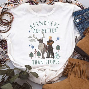 May include: A white short-sleeved t-shirt featuring a graphic print of a reindeer and a person standing together. The text "REINDEERS ARE BETTER THAN PEOPLE" is printed in green above and below the characters, surrounded by small trees and snowflakes.