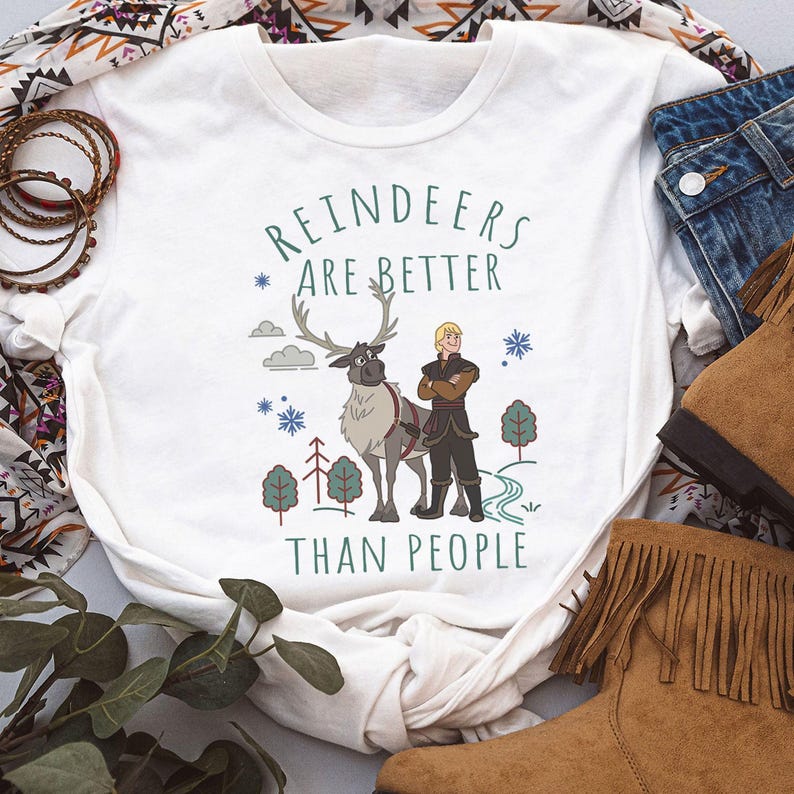 May include: A white short-sleeved t-shirt featuring a graphic print of a reindeer and a person standing together. The text "REINDEERS ARE BETTER THAN PEOPLE" is printed in green above and below the characters, surrounded by small trees and snowflakes.