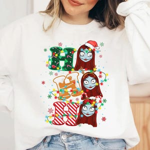 Peut inclure: Sweat-shirt blanc avec un motif de No&euml;l. Le texte "HO HO HO" est compos&eacute; d'&eacute;l&eacute;ments festifs : vert avec des lumi&egrave;res, pain d'&eacute;pices et rayures rouges et blanches. Sally de L'&Eacute;trange No&euml;l de Monsieur Jack est repr&eacute;sent&eacute;e.