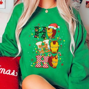 May include: Green sweatshirt with a festive "Ho Ho Ho" design. The design features Iron Man's helmet incorporated into the letters, with Christmas lights and a Santa hat. The sweatshirt is a vibrant green color.
