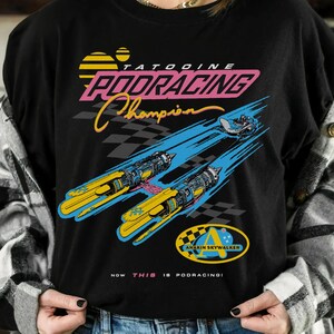 以下が含まれることがあります： レトロなグラフィックデザインの黒いTシャツ。「Tatooine Podracing Champion」の文字と、黄色と青のポッドレーサー、チェッカーフラッグが描かれています。デザインには「Anakin Skywalker」の文字も含まれています。ヴィンテージ感のあるTシャツです。