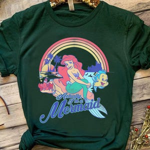 Pode incluir: T-shirt verde escuro com um gr&aacute;fico colorido de uma sereia, um arco-&iacute;ris e o texto "Actually I'm a Mermaid". O design inclui uma sereia com cabelo ruivo e um peixe. A camisa tem mangas curtas e gola redonda.