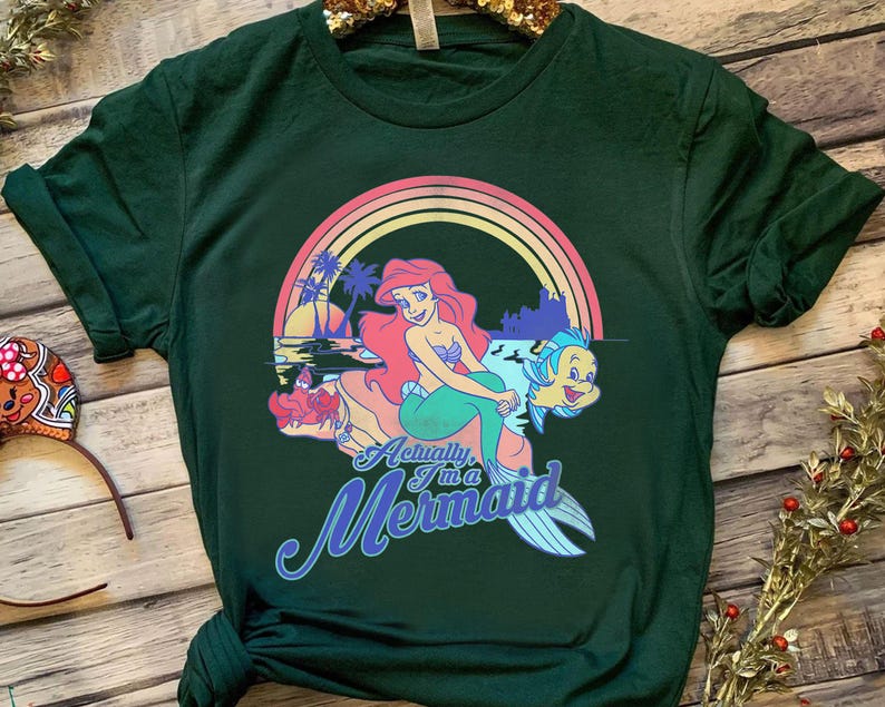 Pode incluir: T-shirt verde escuro com um gr&aacute;fico colorido de uma sereia, um arco-&iacute;ris e o texto "Actually I'm a Mermaid". O design inclui uma sereia com cabelo ruivo e um peixe. A camisa tem mangas curtas e gola redonda.