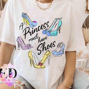 May include: A white short-sleeve t-shirt featuring a graphic print of five colorful high-heeled shoes and the black text A Princess must have Shoes. The shoes are illustrated in blue, yellow, and purple with names written on each shoe.