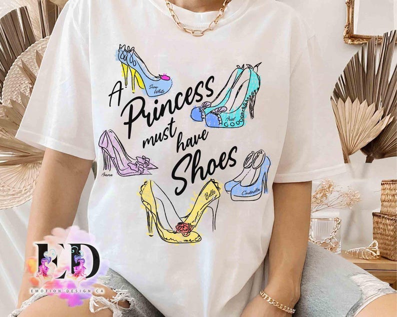 May include: A white short-sleeve t-shirt featuring a graphic print of five colorful high-heeled shoes and the black text A Princess must have Shoes. The shoes are illustrated in blue, yellow, and purple with names written on each shoe.