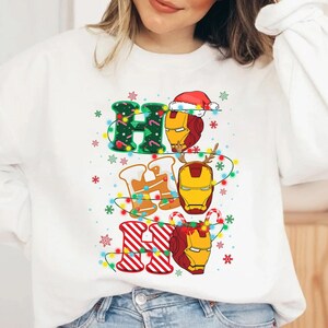 May include: White sweatshirt with a festive "HO HO HO" design. The letters are decorated with Christmas lights, snowflakes, and Iron Man's helmet. The "H" is green with a Christmas tree pattern, the "O" has a Santa hat, and the last "HO" is red and white striped.