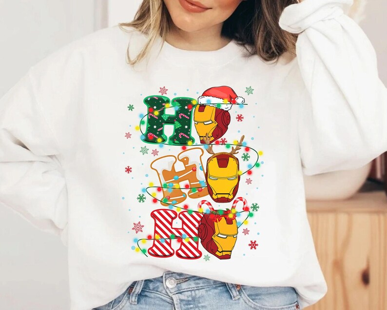 May include: White sweatshirt with a festive "HO HO HO" design. The letters are decorated with Christmas lights, snowflakes, and Iron Man's helmet. The "H" is green with a Christmas tree pattern, the "O" has a Santa hat, and the last "HO" is red and white striped.